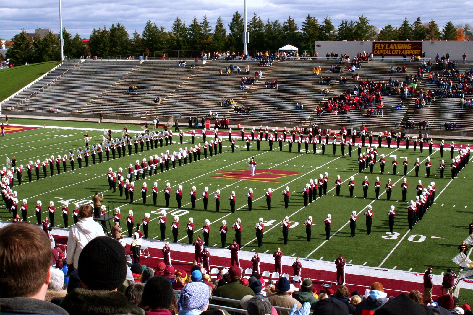 A college marching band in maroon and white uniforms forms a large open C on a football field at halftime, with partly filled grandstands and a crowd in the foreground.
