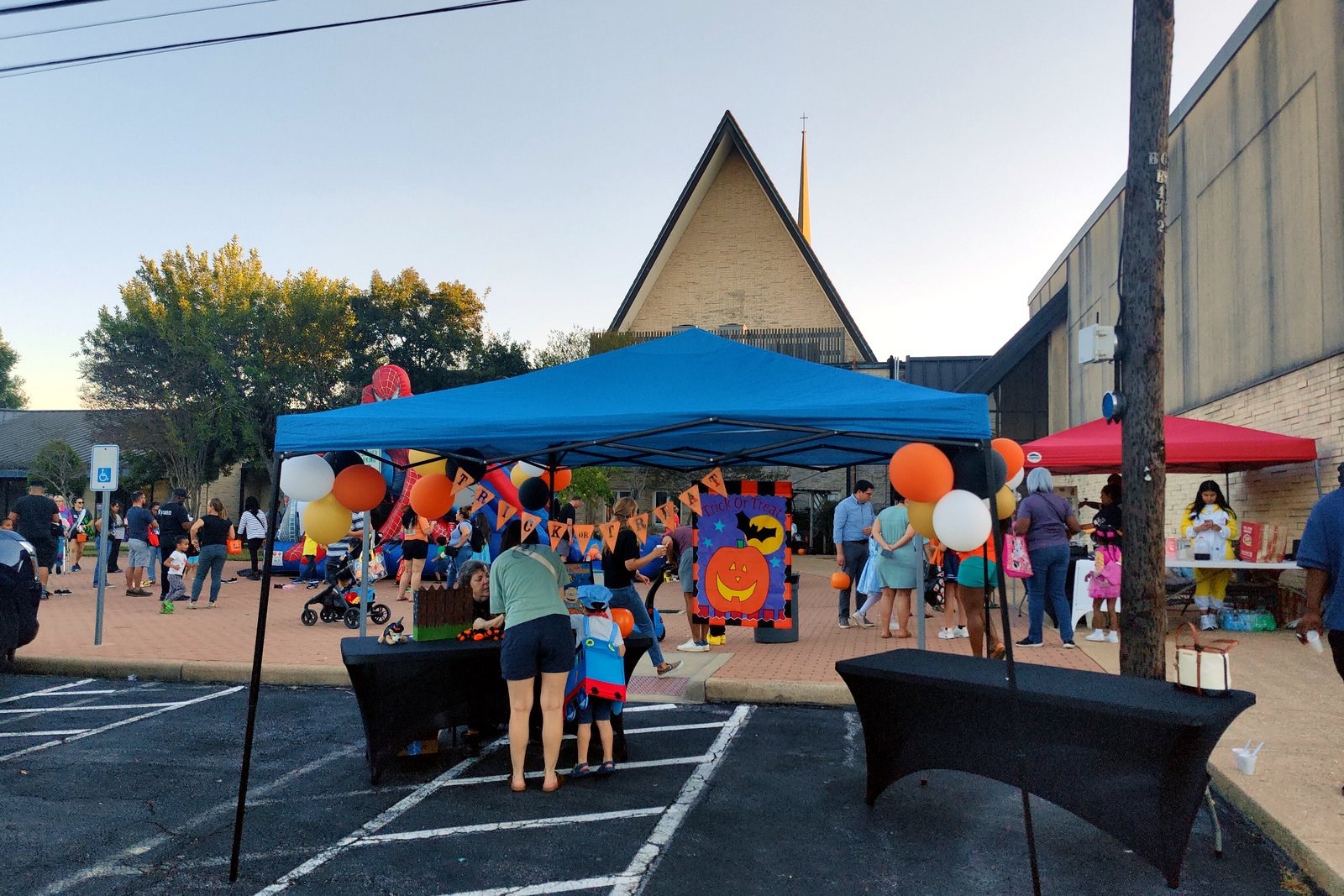 Families gather under a canopy tent decorated with orange and white balloons at an outdoor trunk-or-treat in a church parking lot.