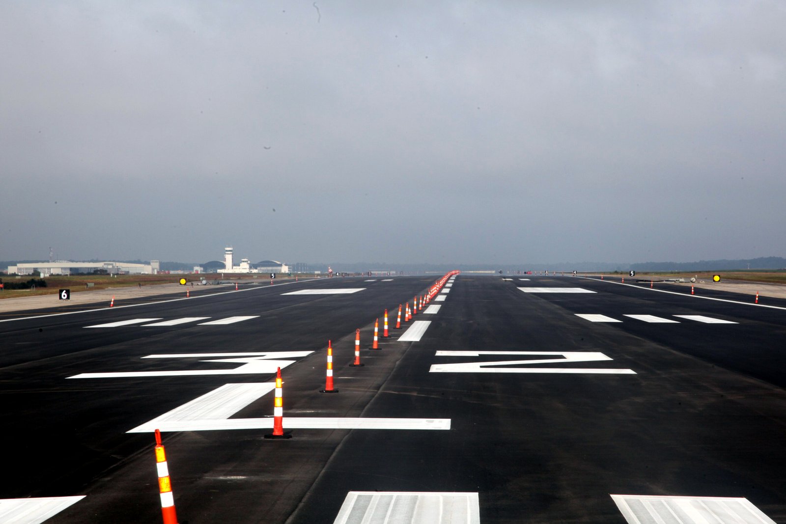 A freshly paved dark asphalt runway with bright white markings and a line of orange-and-white traffic cones down its center, stretching toward a distant control tower under a gray sky.