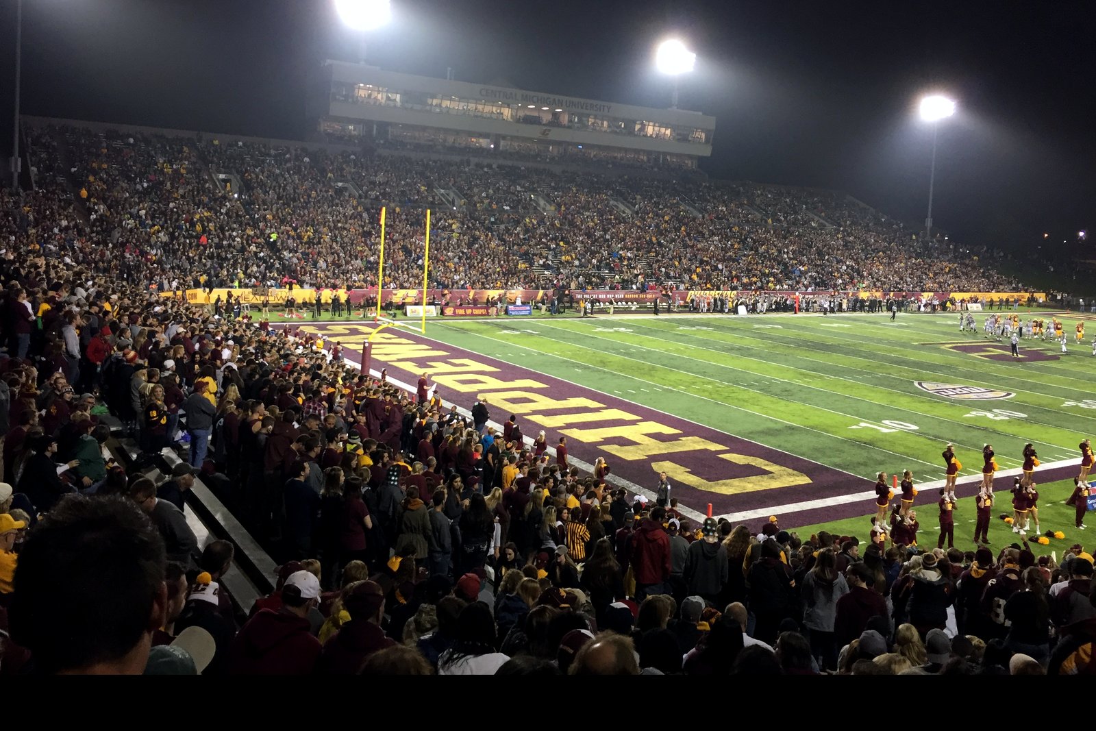 A packed football stadium at night under bright light towers, with fans in maroon and gold in the foreground and a maroon end zone lettered in gold.