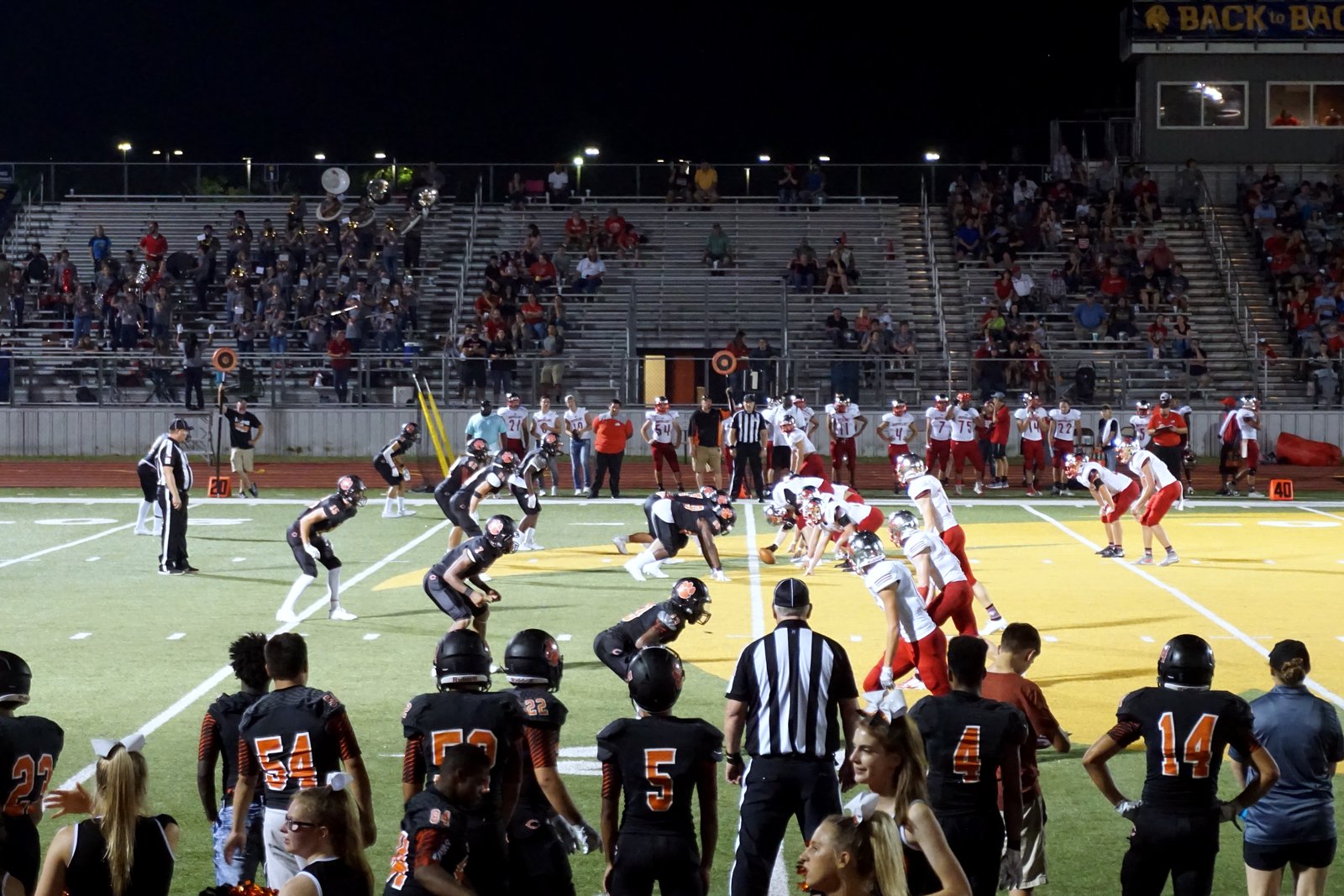 High school football players in white and black jerseys line up on a field at night as teammates watch from the sideline in front of crowded stands.
