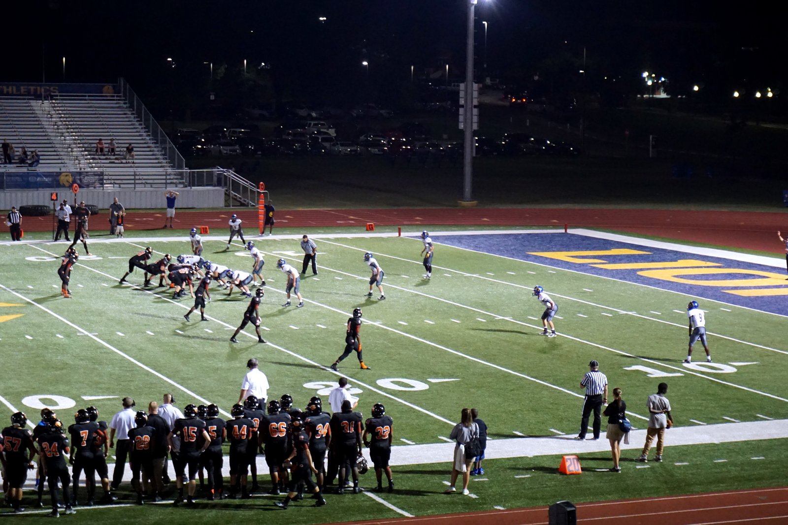 Football players in dark uniforms line up on offense against a team in white on a grass field, with a packed sideline and stadium stands behind them.