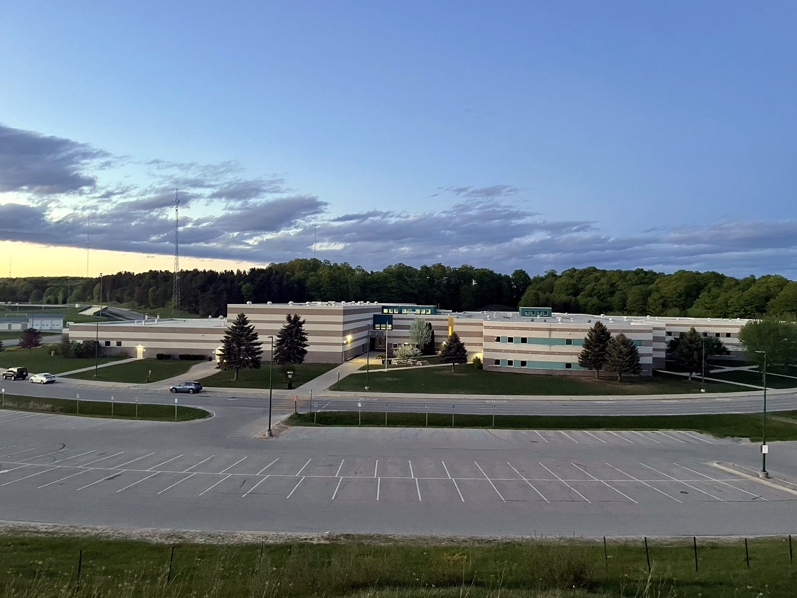 A long, low school building stretches across a tree line beyond an empty parking lot and lawns under a blue evening sky.
