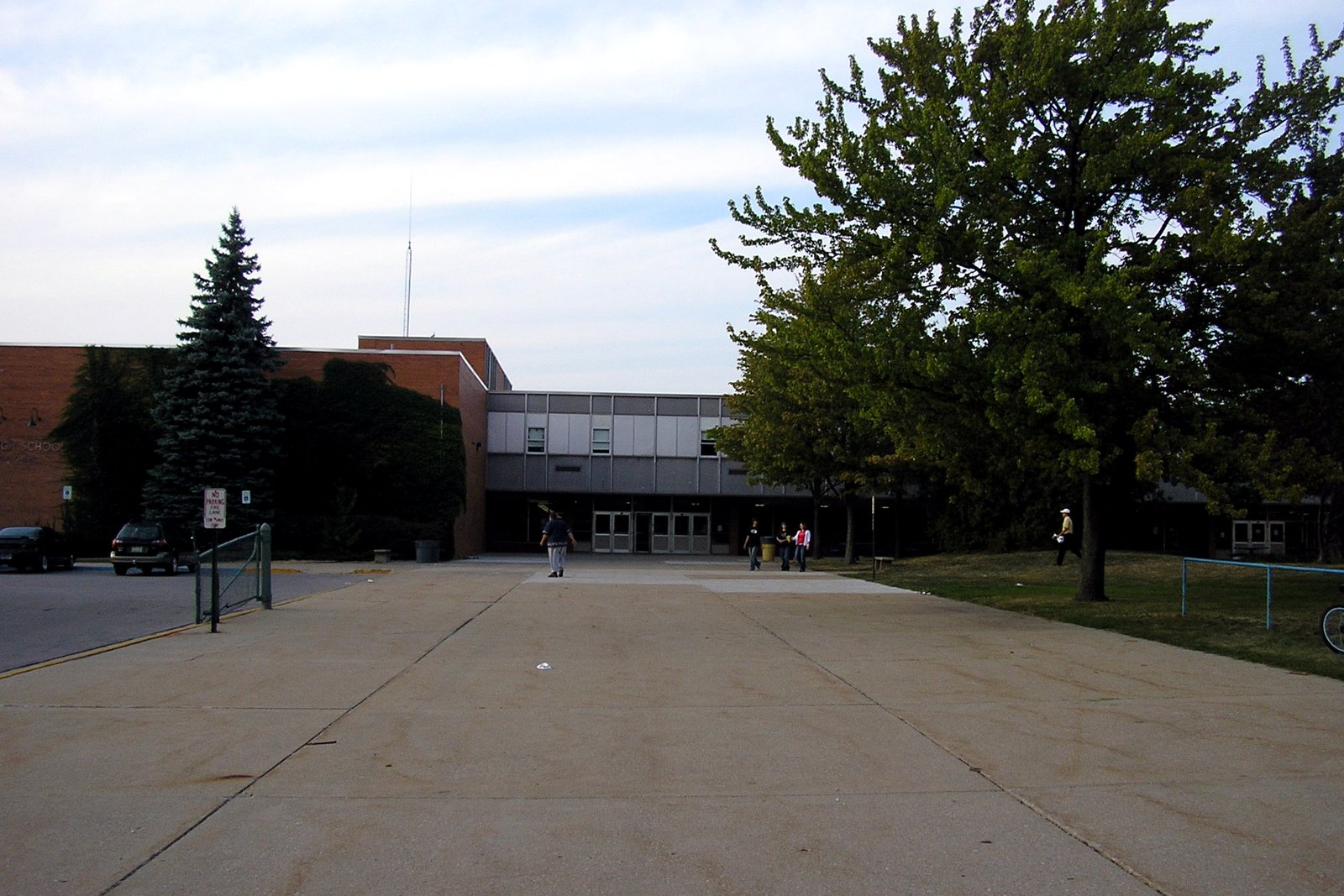 A wide concrete walkway leads to the entrance of a brick and gray-paneled school building, with a few people near the doors, an evergreen tree at left and leafy trees at right.