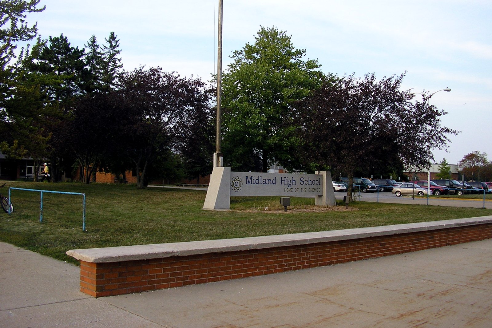 A low stone sign reading Midland High School on a lawn beside a flagpole and trees, with a curved brick planter wall and a parking lot behind it.