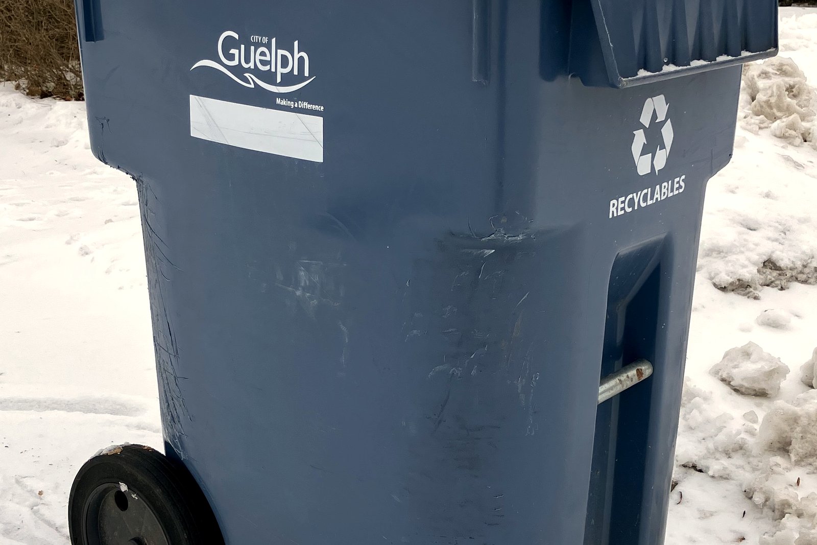 Close-up of a blue wheeled recycling cart with a recycling symbol and the word Recyclables, standing beside snow.