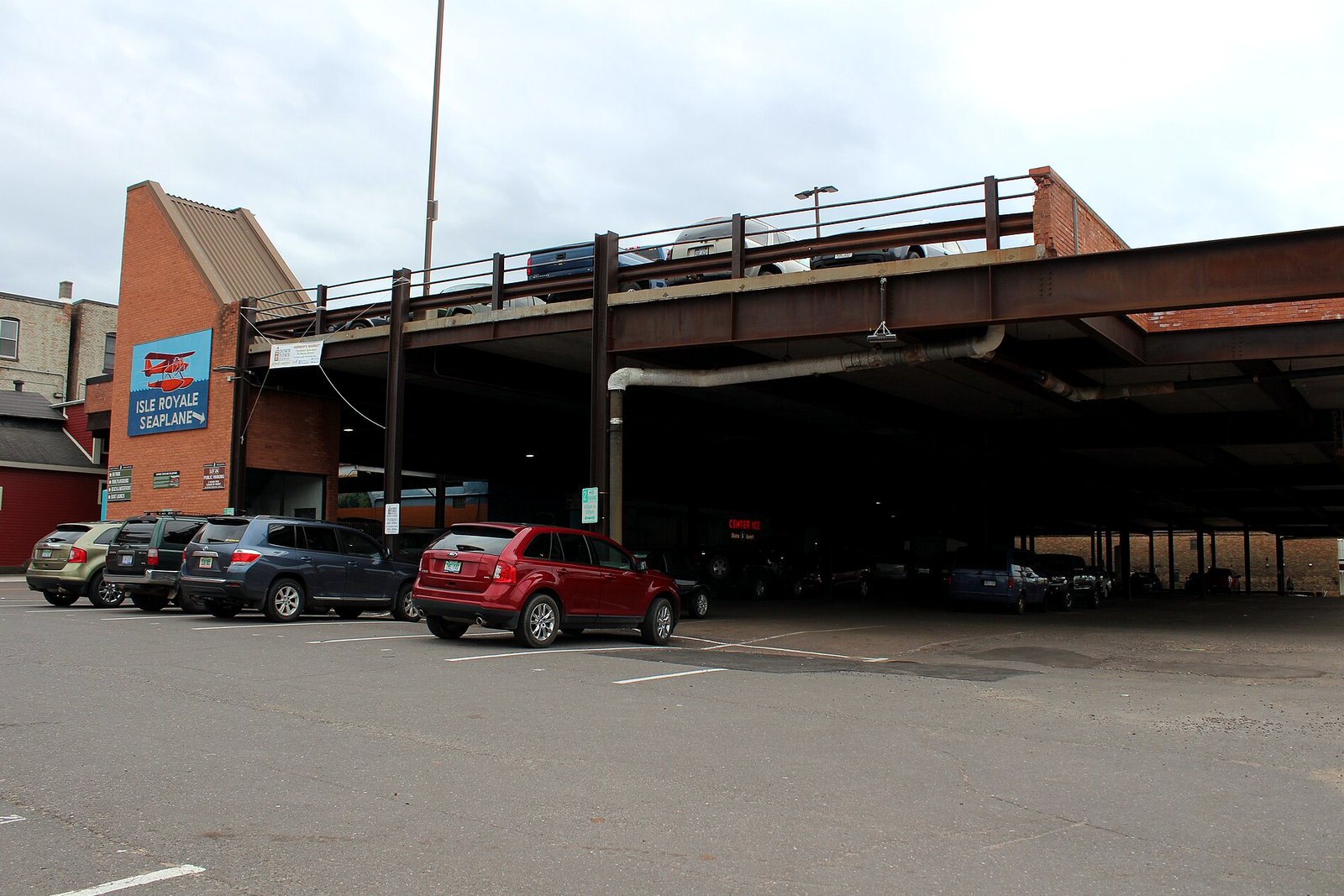 A steel-and-concrete parking deck with cars parked beneath it and on its top level, beside brick buildings under a cloudy sky.