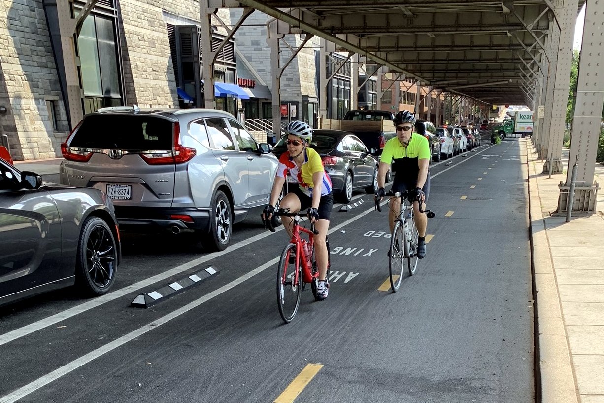 Two cyclists ride in a two-way bike lane separated from traffic by a row of parked cars, beneath an elevated structure on a city street.