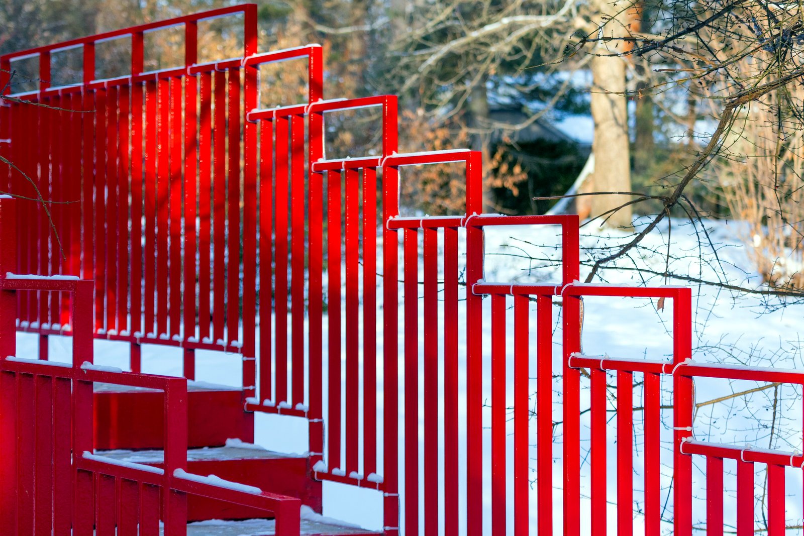 Bright red metal railings of a footbridge crossing a snowy garden, with bare trees behind.