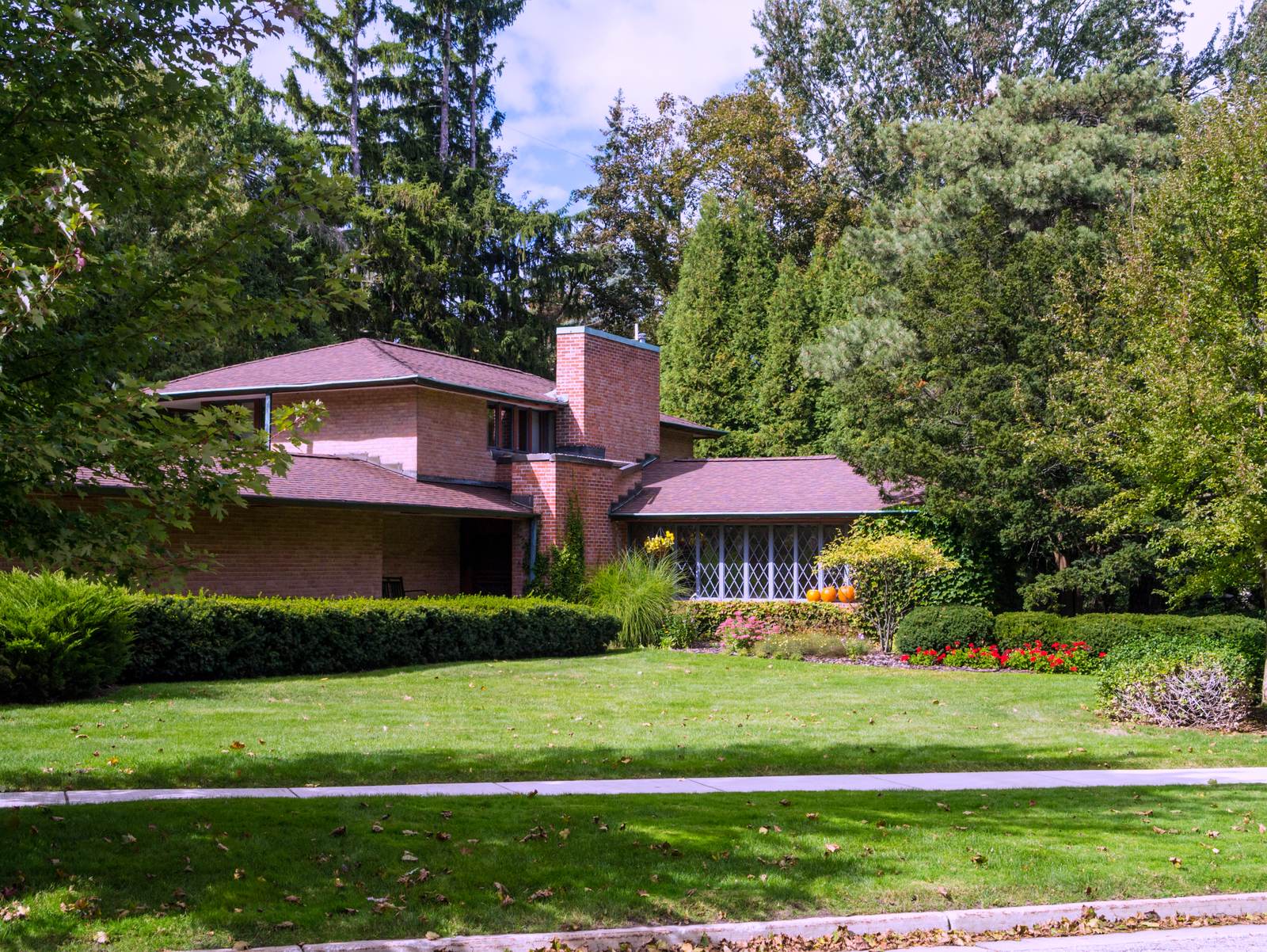 A low brick house with a red-brown roof behind a wide green lawn, framed by tall trees.