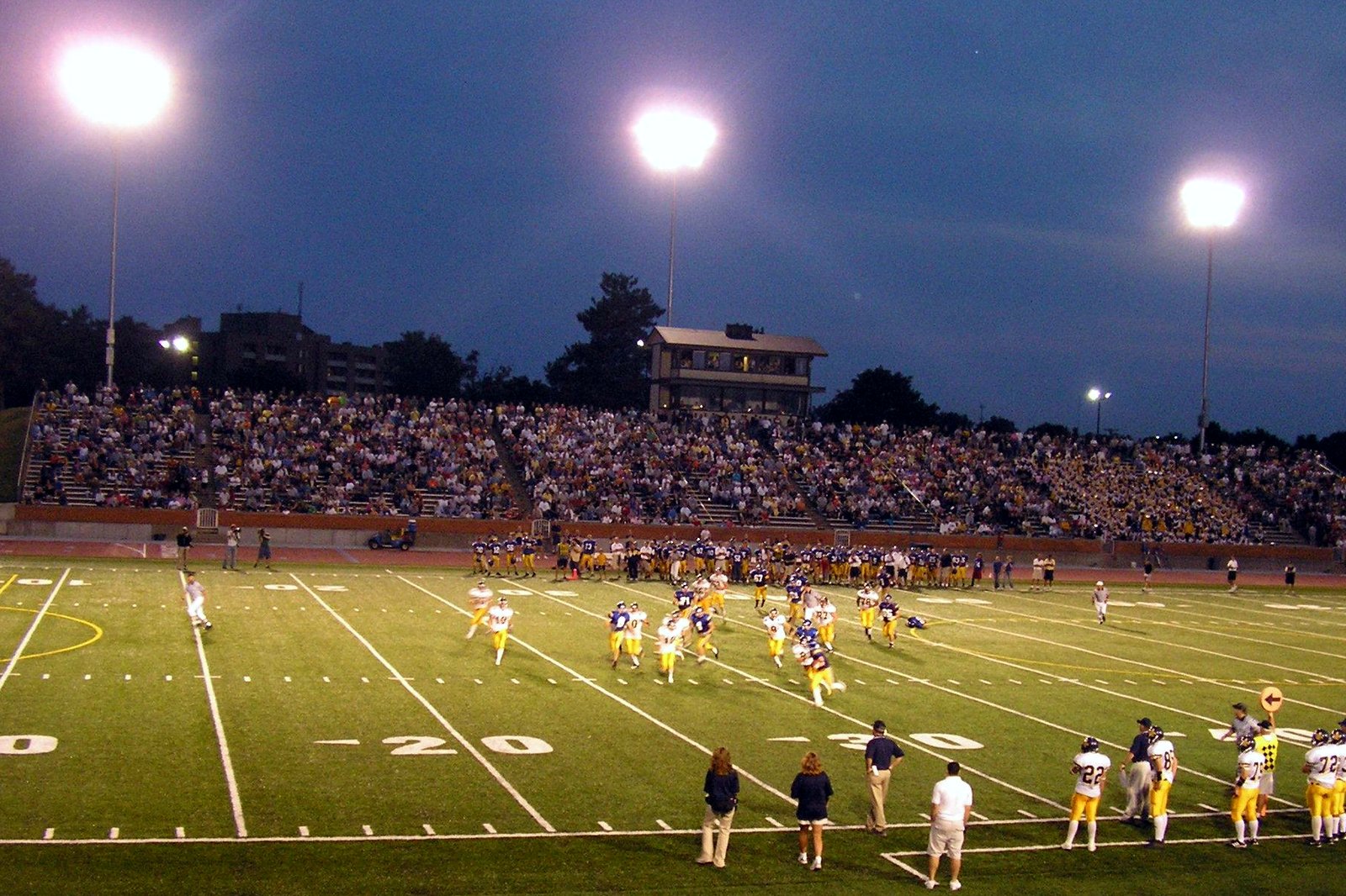 A high school football game at night under bright stadium lights, with players in gold and white on a green field and a crowded grandstand behind them.