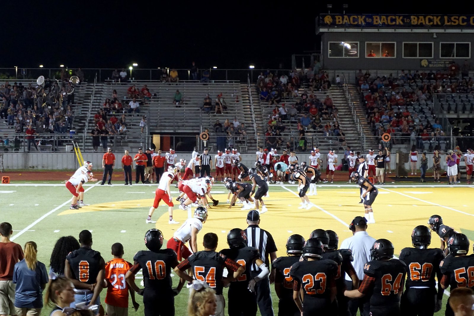 High school football players lined up at the line of scrimmage during a daytime game, with fans in the stands behind them.