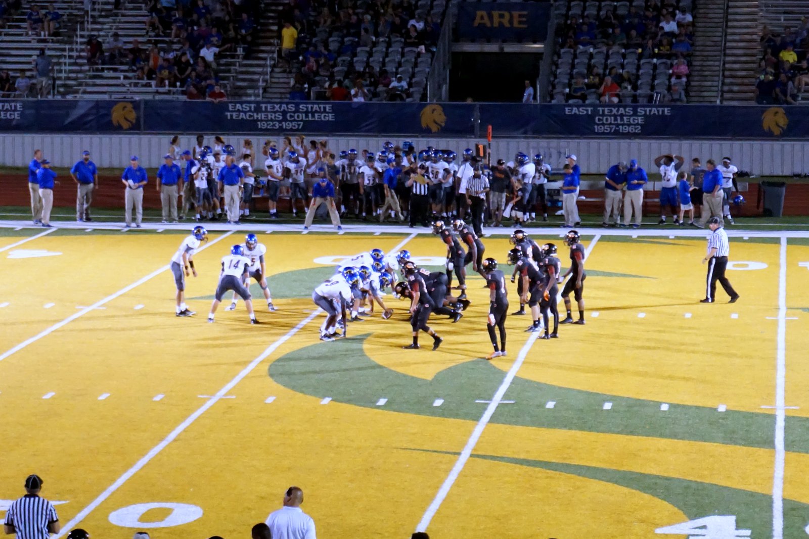 High school football players in dark and light uniforms line up at the line of scrimmage under stadium lights.