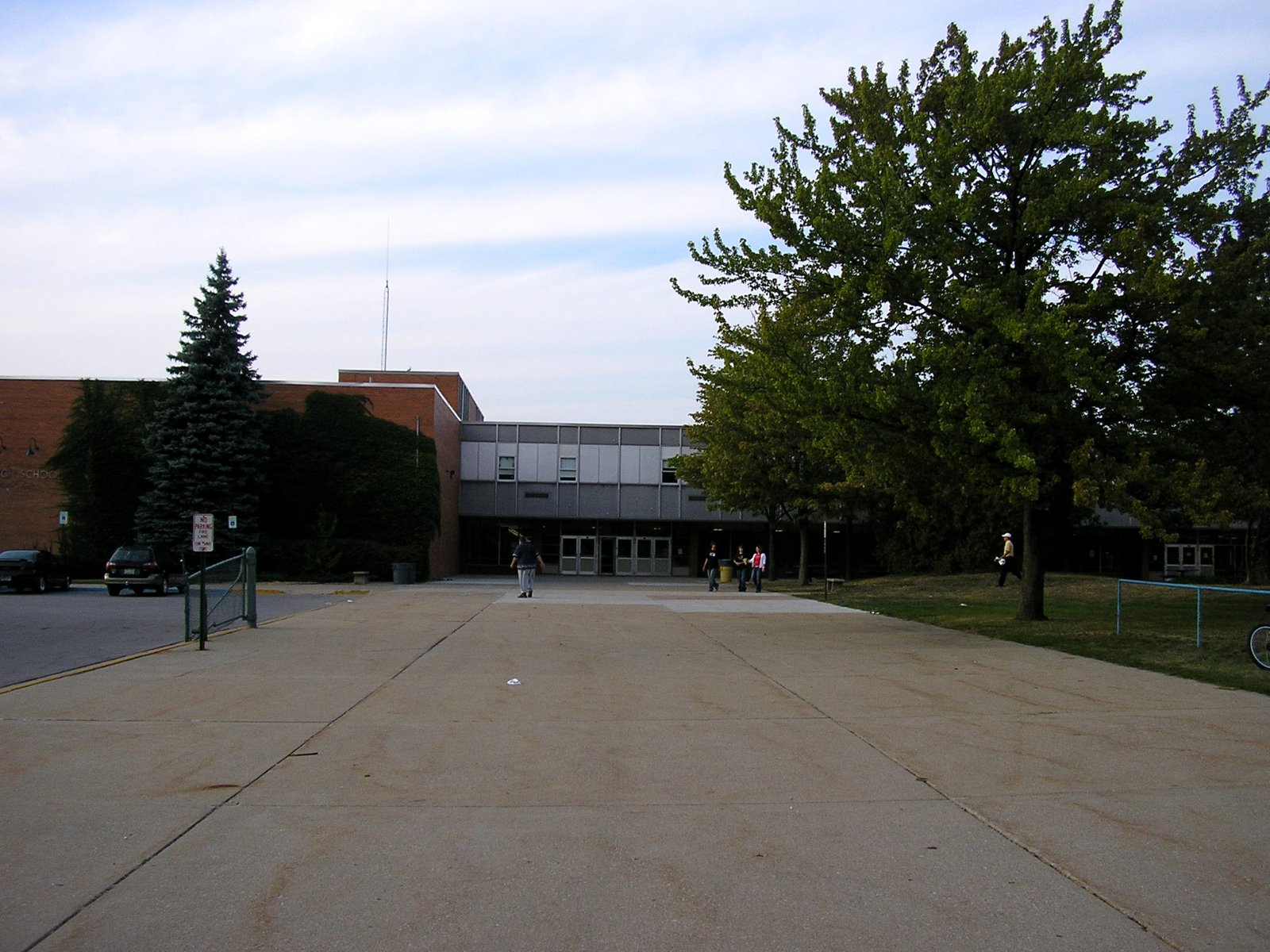 A long brick and gray-paneled school building with glass entry doors at the end of a wide concrete plaza, with a tall evergreen at left and a leafy tree at right under a pale sky.
