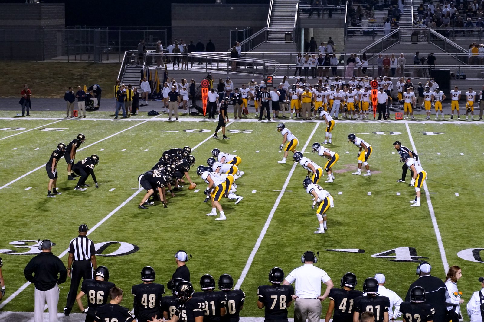 Players in white and gold line up on offense against a team in dark jerseys on a turf field, with players and officials along the sideline.