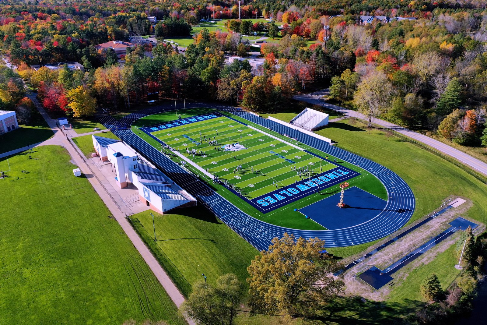 Aerial view of a football field with blue end zones reading Northwood and Timberwolves, ringed by a blue running track and surrounded by trees in fall color.