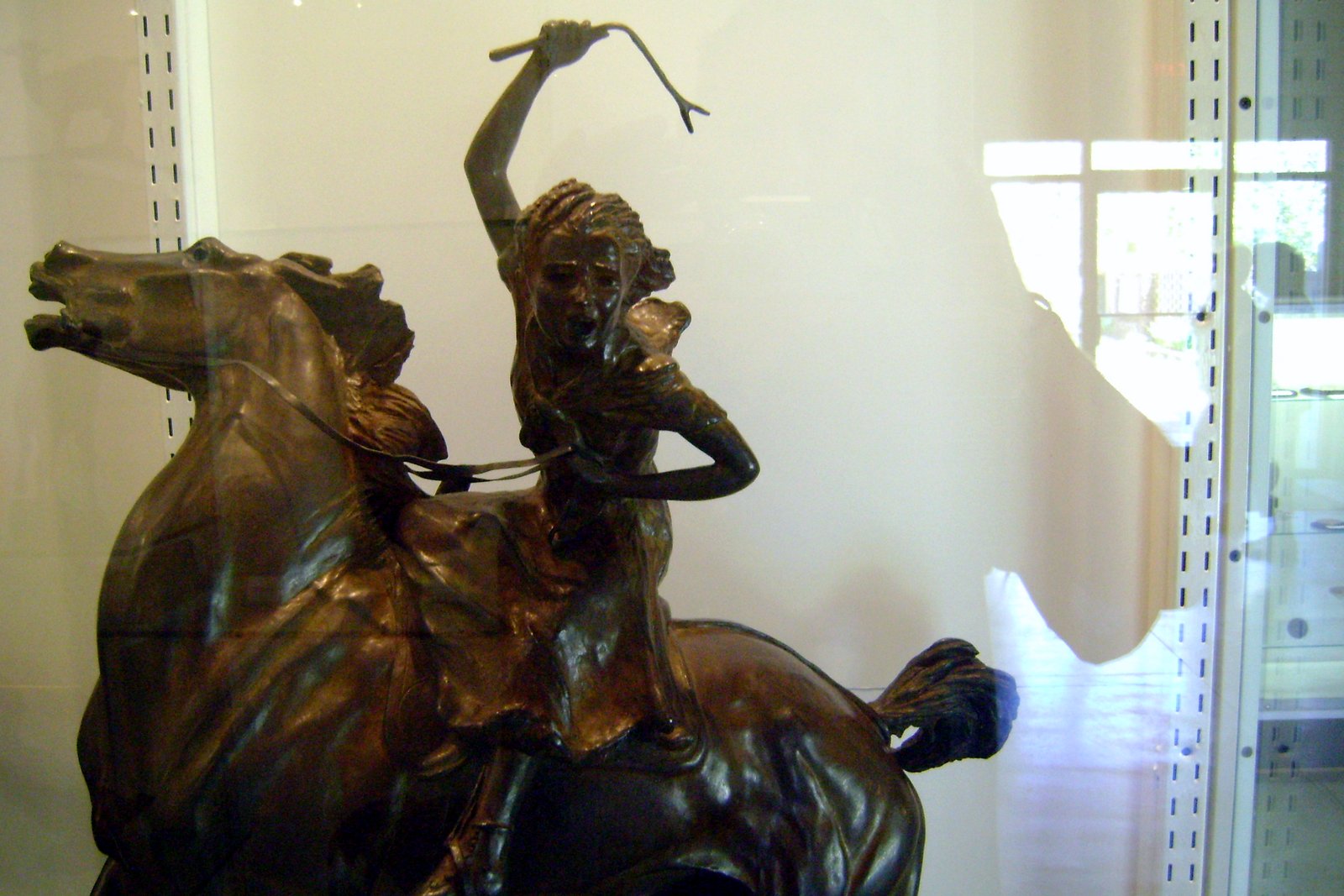 A bronze sculpture of a young woman on a rearing horse, one arm raised high holding a stick, displayed in a glass case.