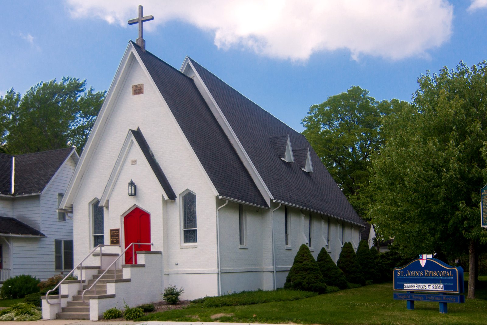 A white-painted brick church with a steep gray roof, a cross on its peak, red front doors at the top of a staircase and a blue St. John’s Episcopal sign on the lawn.