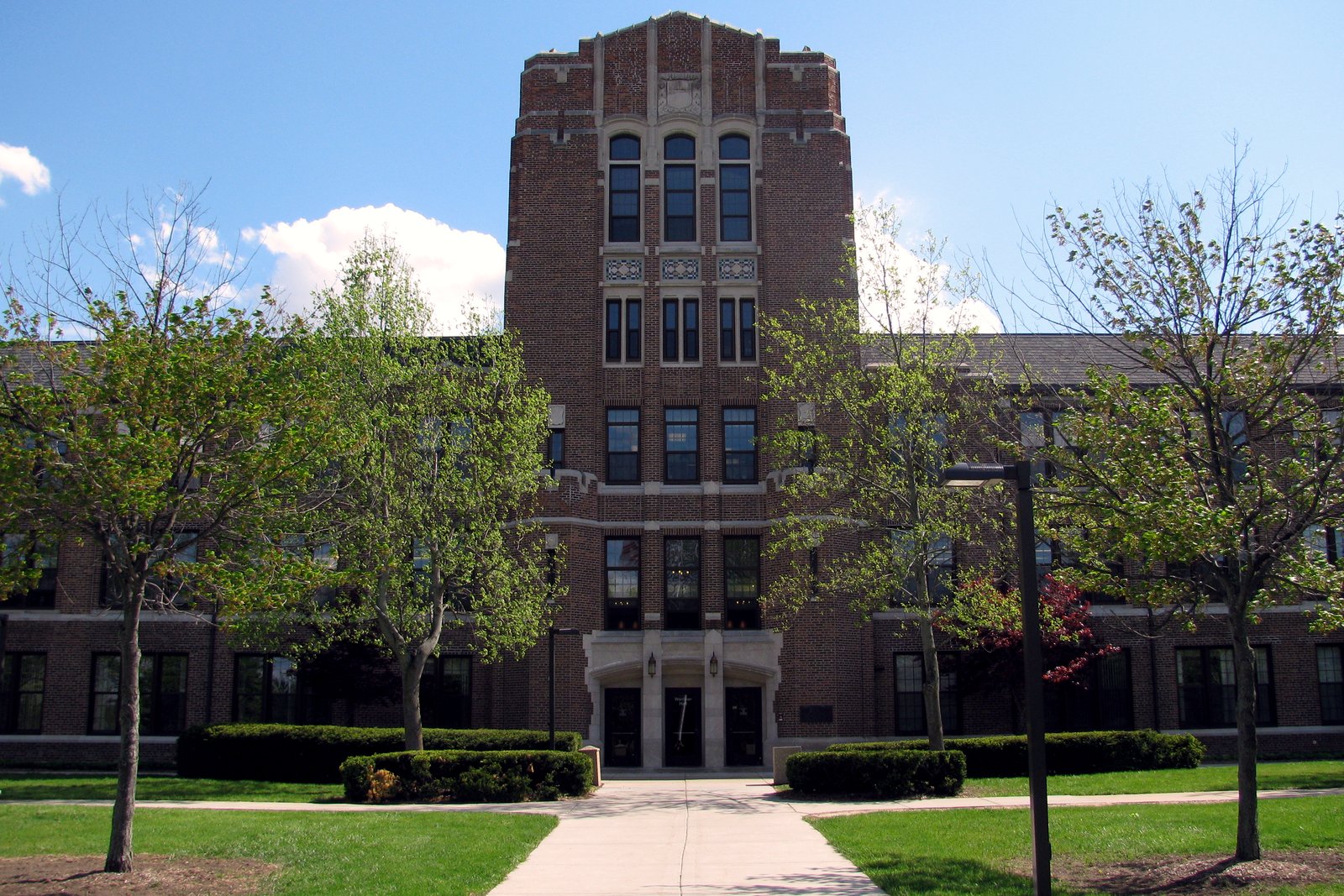 A tall brick academic building with a central tower and arched entrance, behind a green lawn and trees.