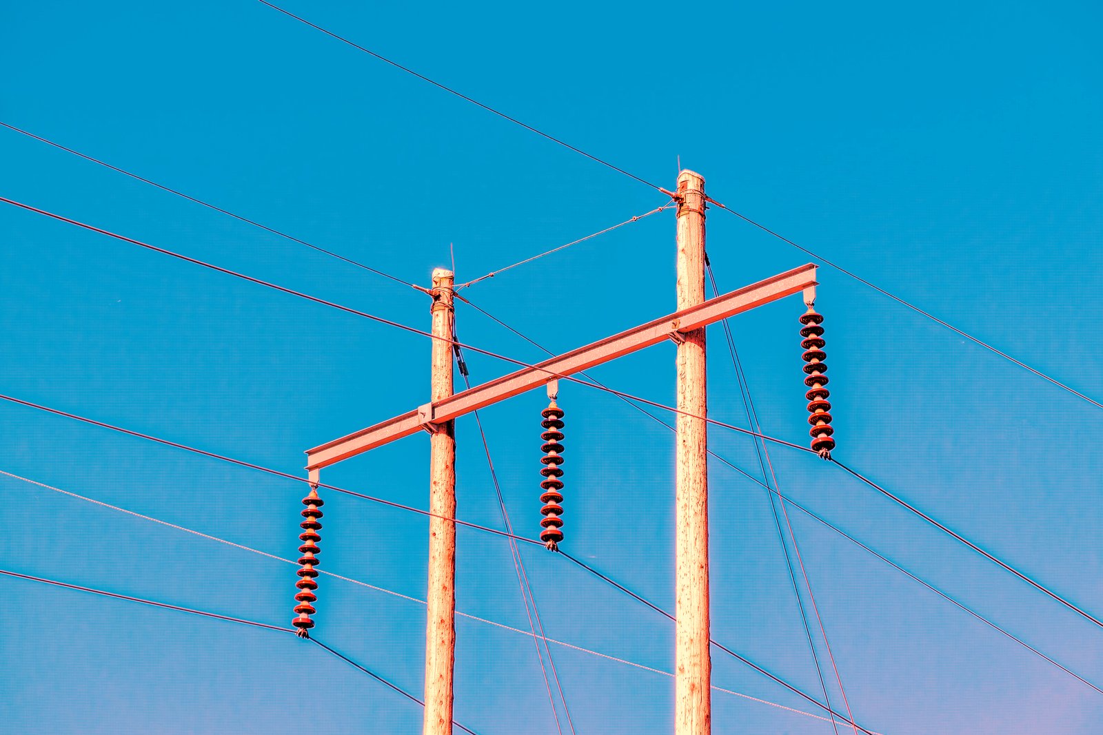 Two wooden utility poles joined by a metal crossarm with hanging insulators and power lines against a clear blue sky.