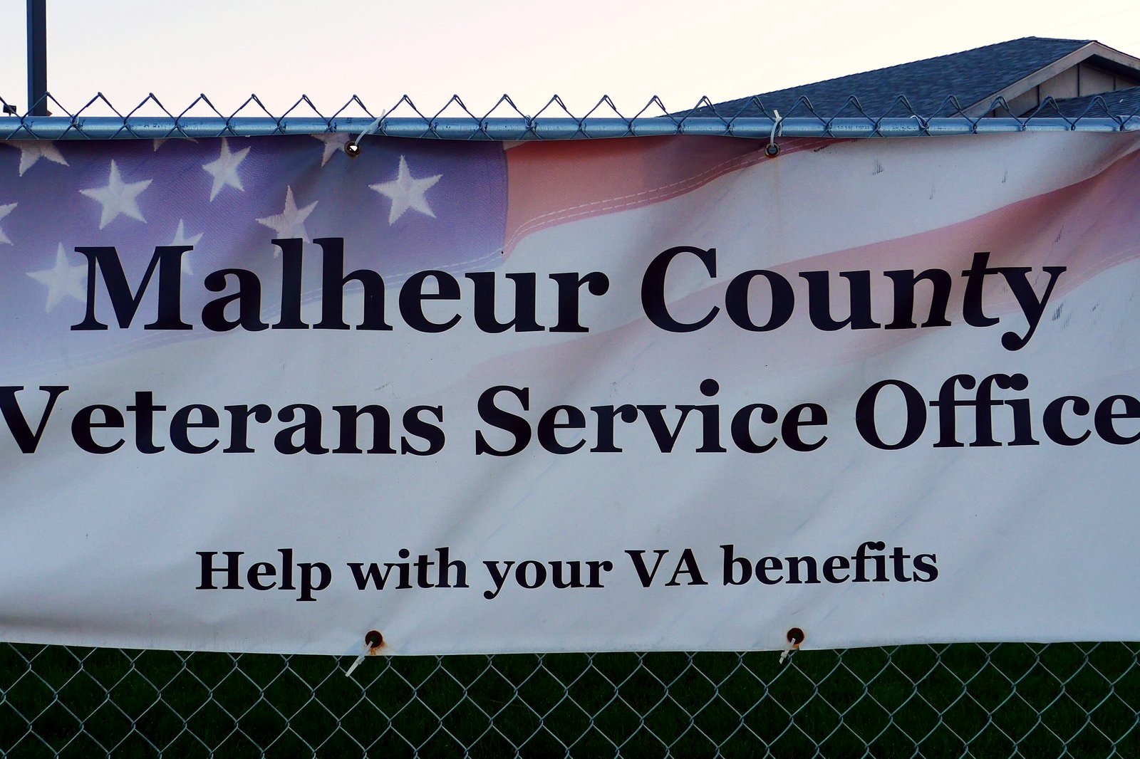 A white vinyl banner with a faded American flag design, zip-tied to a chain-link fence, reading Malheur County Veterans Service Office, Help with your VA benefits.