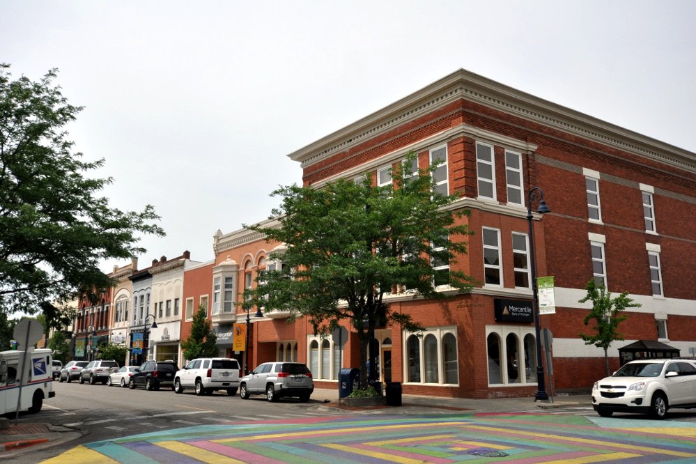 A row of two- and three-story brick commercial buildings on a downtown corner with parked cars and a painted crosswalk.