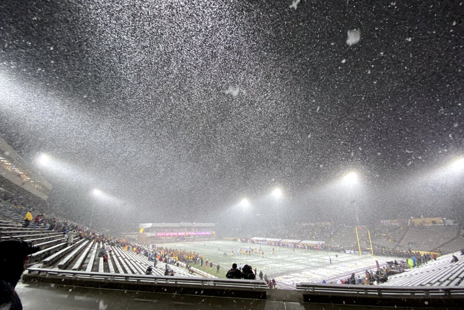 A football stadium at night under heavy falling snow, with players on the field and lights glaring over the stands.