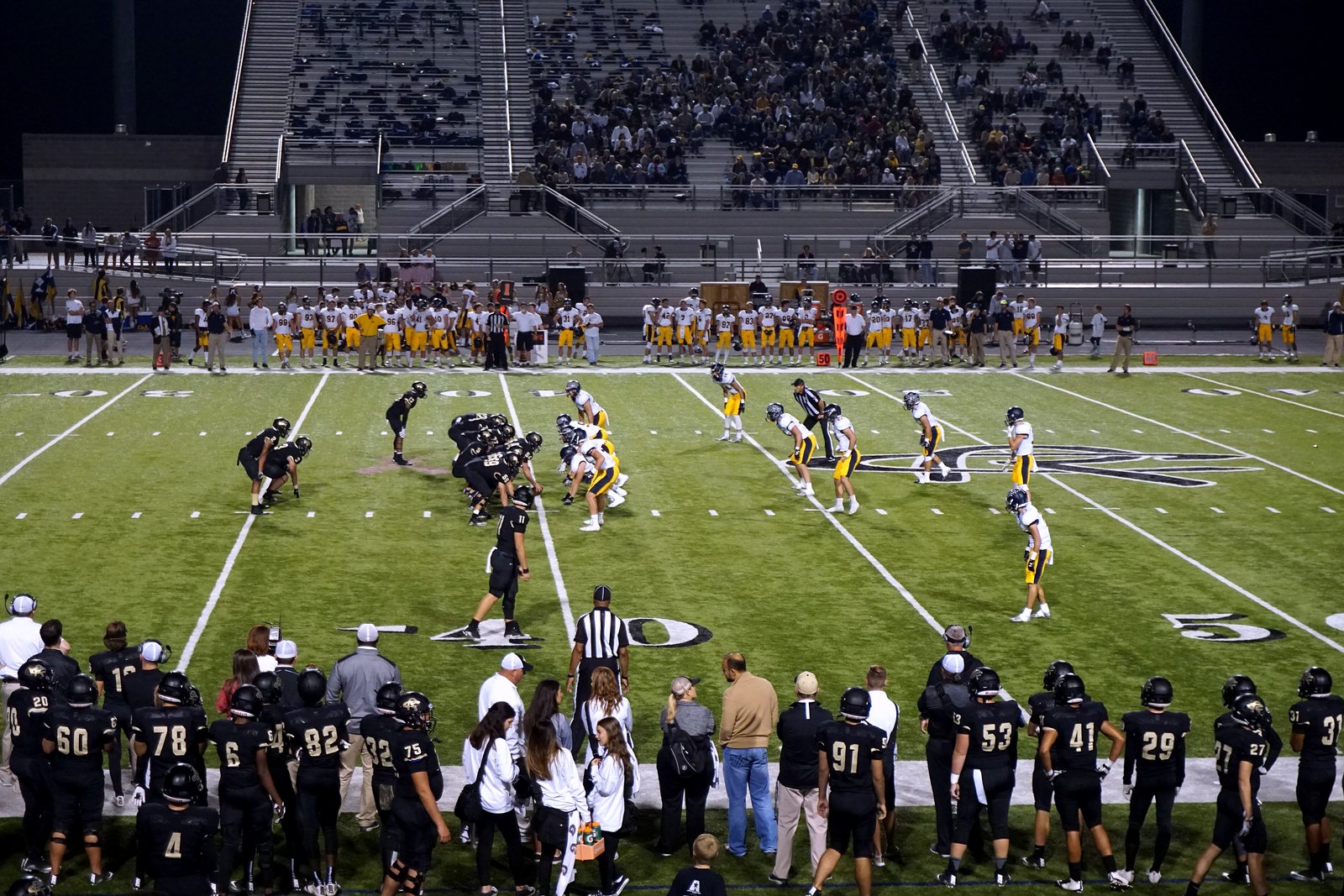 High school football players in red and white uniforms run a play on a green field, with a crowd in the stands.