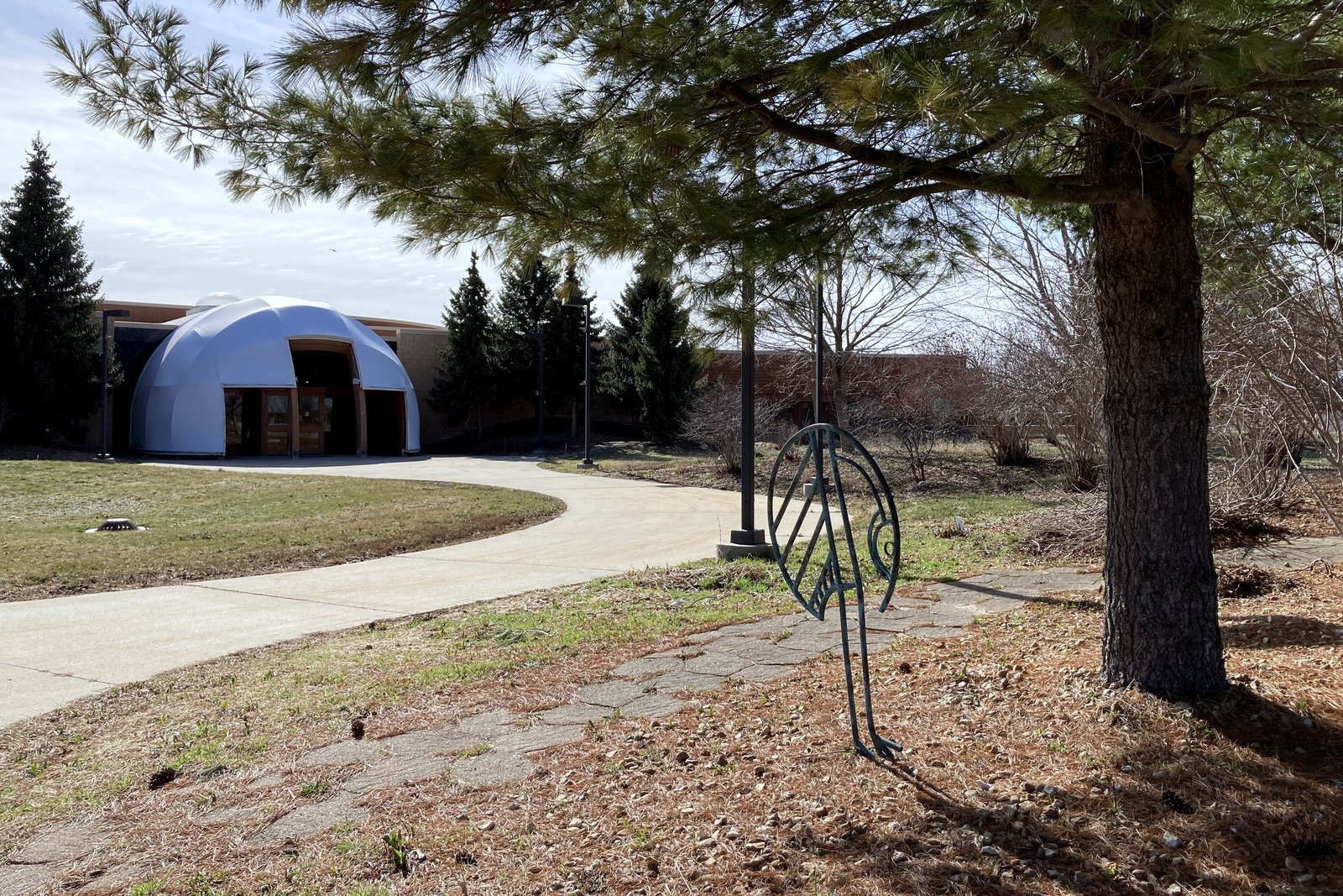 A paved path curving across a lawn toward a low domed building, with a pine tree and a metal sculpture in the foreground.