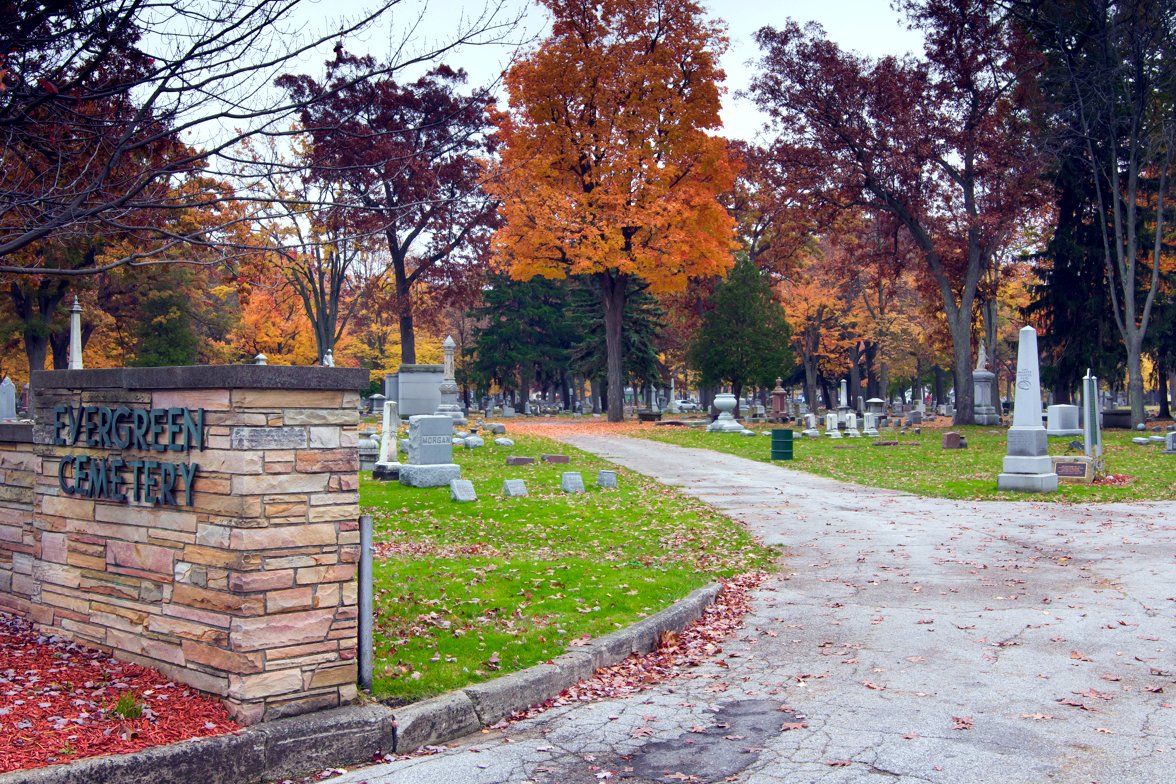 A stone entrance sign reading Evergreen Cemetery stands beside a curving paved drive lined with orange and gold autumn trees and headstones.
