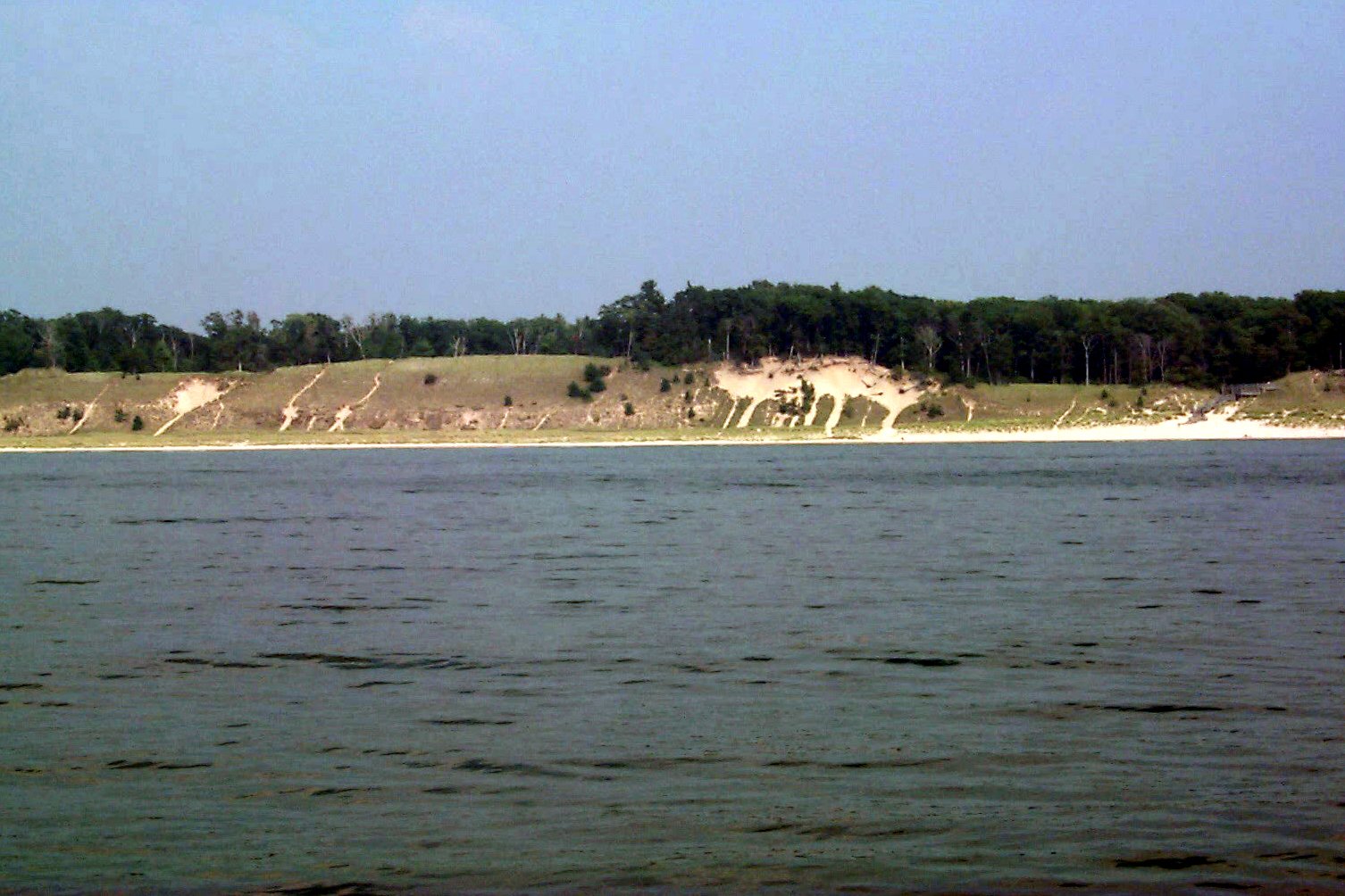 Pale sand dunes topped with trees line the far shore beyond choppy gray-green water along the Muskegon shoreline under a hazy sky.