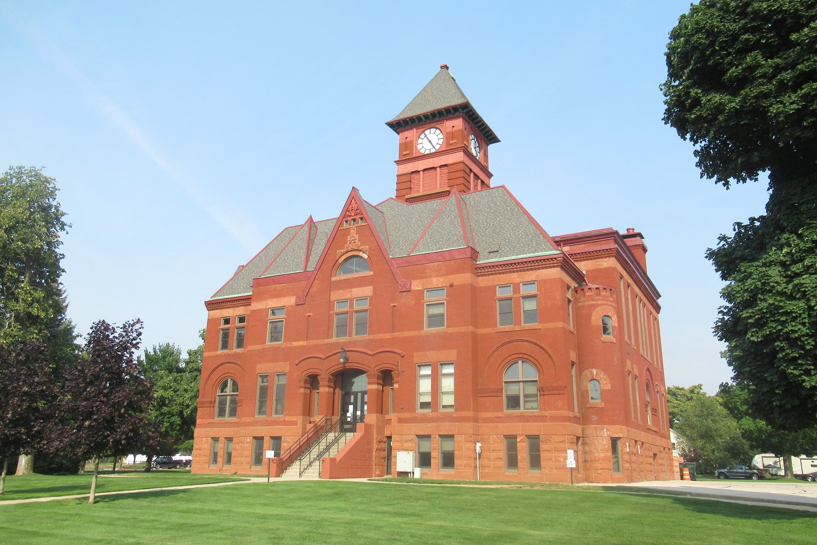 The red sandstone Mason County Courthouse in Ludington, with arched entrance, steep gables and a clock tower, on a sunny day with green lawn and trees.