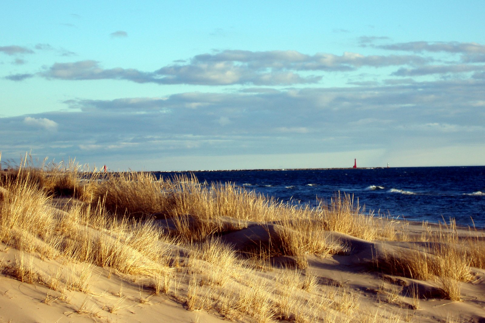 Dry dune grass on a sandy bluff overlooks blue Lake Michigan waves under a cloudy sky, with a channel visible in the distance.