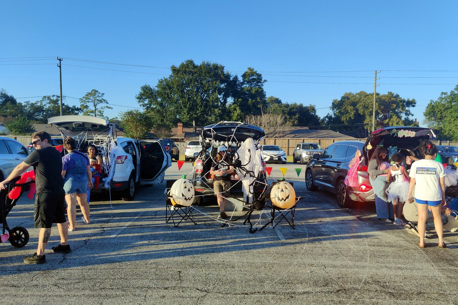 Families walk past decorated car trunks and pumpkin cutouts at a parking-lot trunk-or-treat on a sunny afternoon.