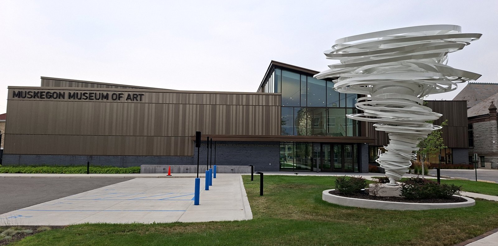 The long, low front of the Muskegon Museum of Art, with its name in large letters on a bronze-colored wall, a glass entrance and a tall white swirling sculpture on the lawn in front.
