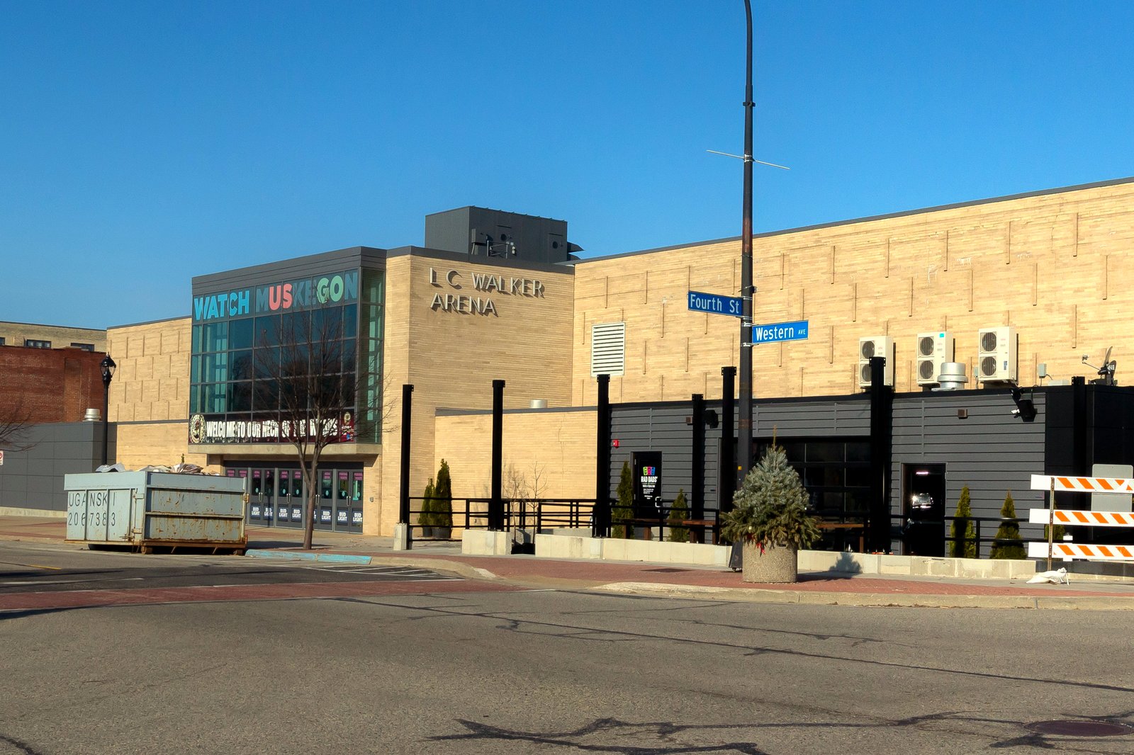 The tan block exterior of the downtown Muskegon hockey arena, with its name in raised letters above a glass entrance and a colorful mural along a lower wall, at a street corner.