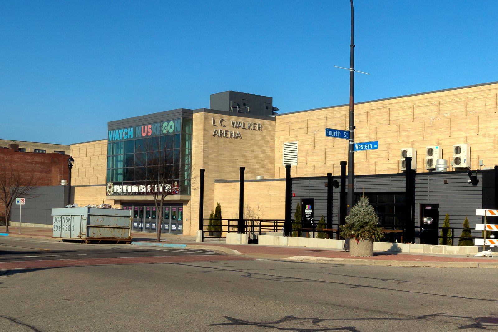 A tan block arena with a glass entrance tower and “L.C. Walker Arena” lettering on a downtown street corner under a clear blue sky.