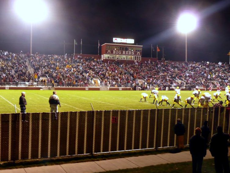 A night football game under bright stadium lights, with a packed grandstand behind a lit field, players in white jerseys lined up, and spectators along a fence in the foreground.