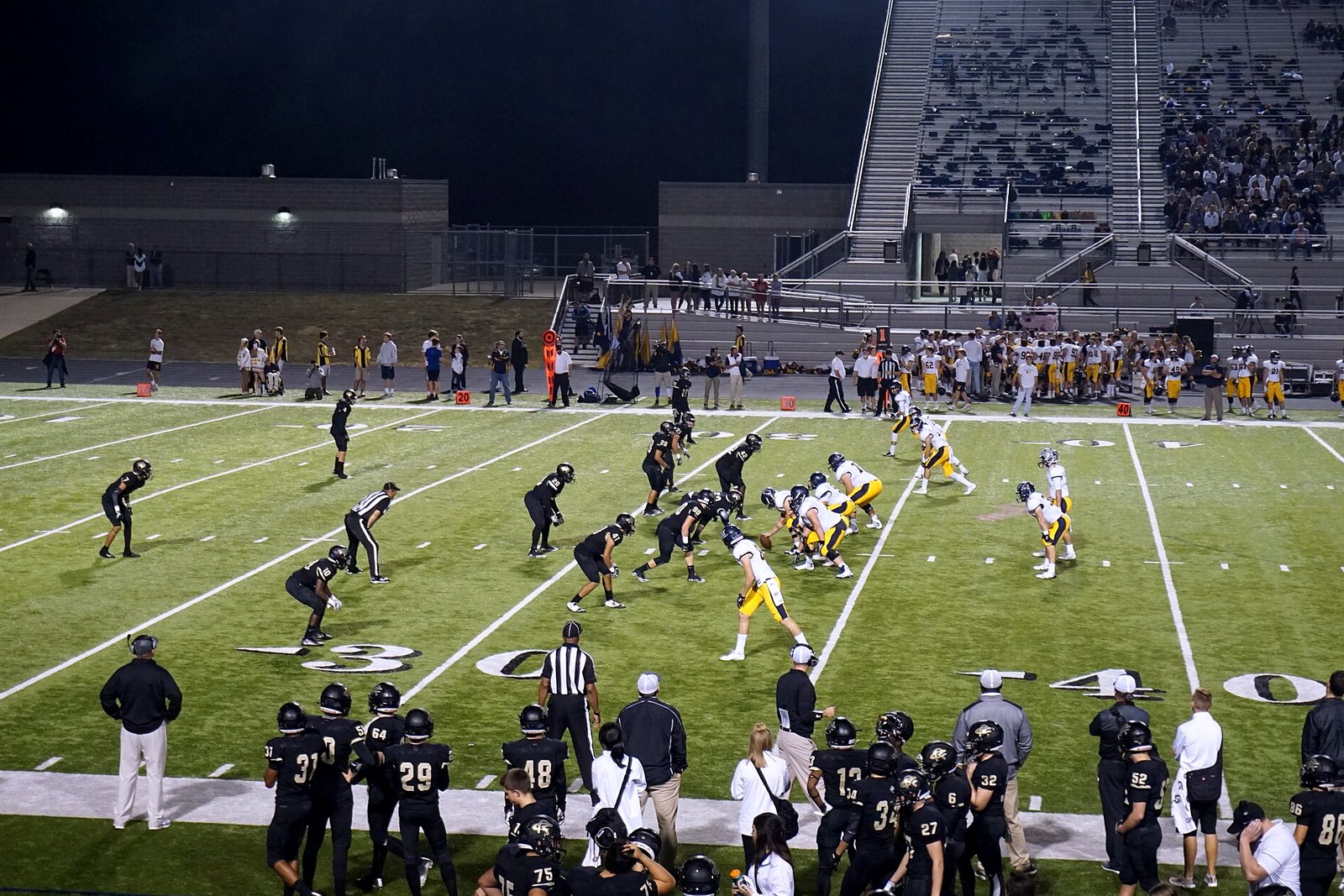 High school football players in dark uniforms run a play on a grass field under stadium lights, with teammates and coaches watching from the sideline.