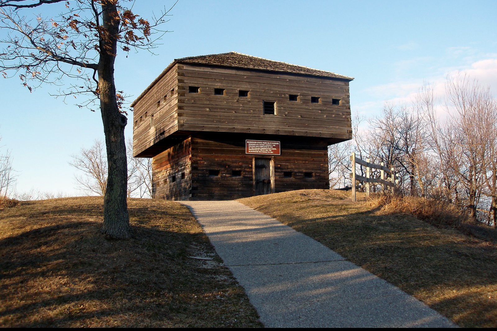 A two-story log blockhouse with small square windows at the top of a grassy hill, reached by a paved walkway, under a clear blue sky in Muskegon State Park.