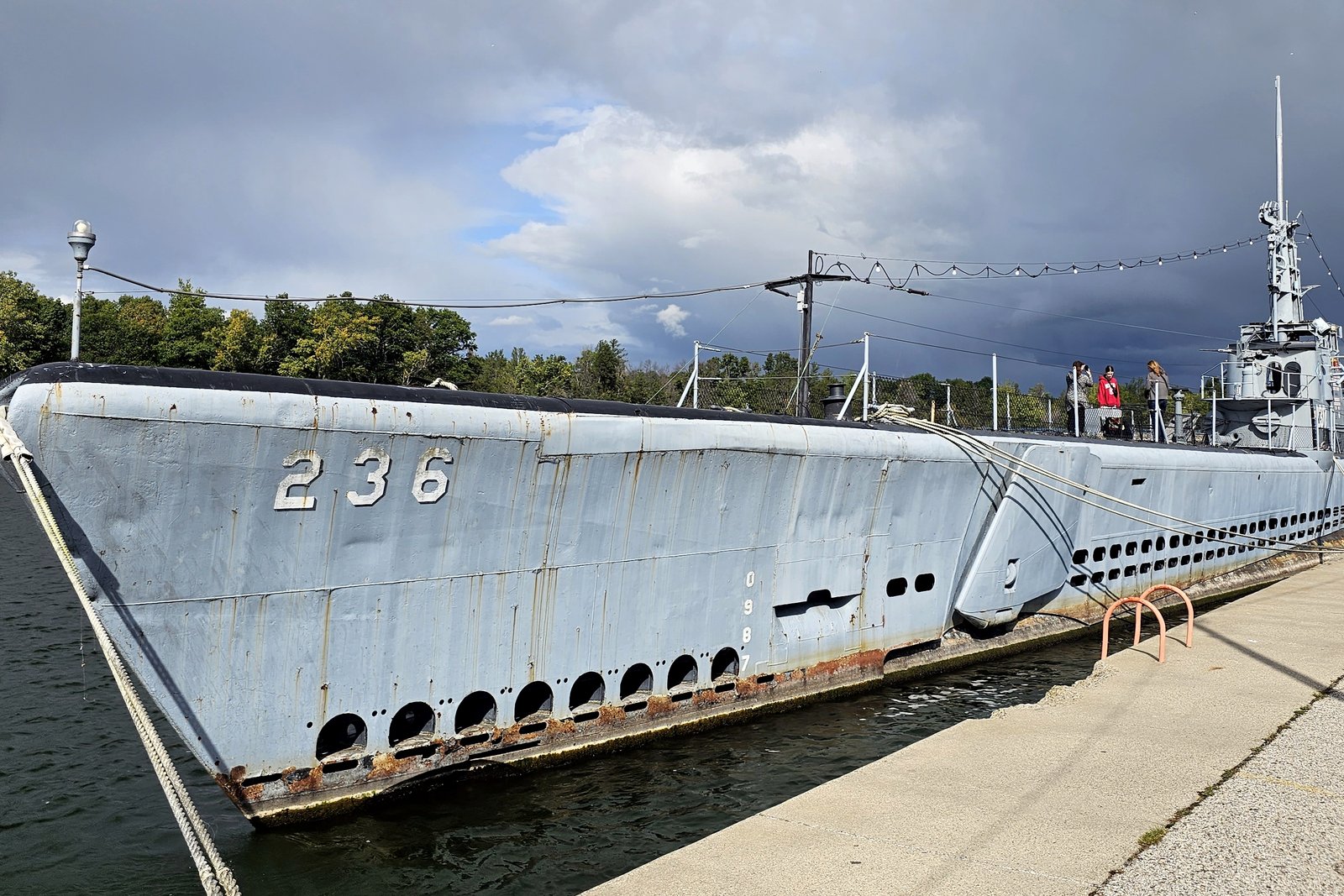 A long gray submarine with the number 236 on its bow moored along a concrete pier, with a tower and antennas amidships under a cloudy sky.