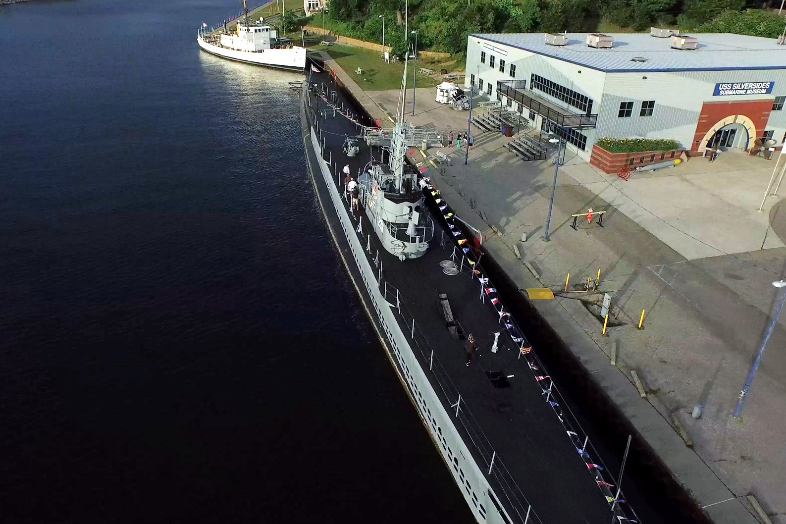 Aerial view of a long gray submarine moored along a concrete pier in a river channel beside a low museum building.