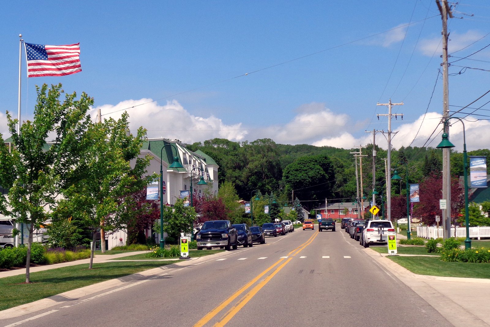 A two-lane village street with green lampposts, an American flag, young trees and small shops under a blue summer sky.