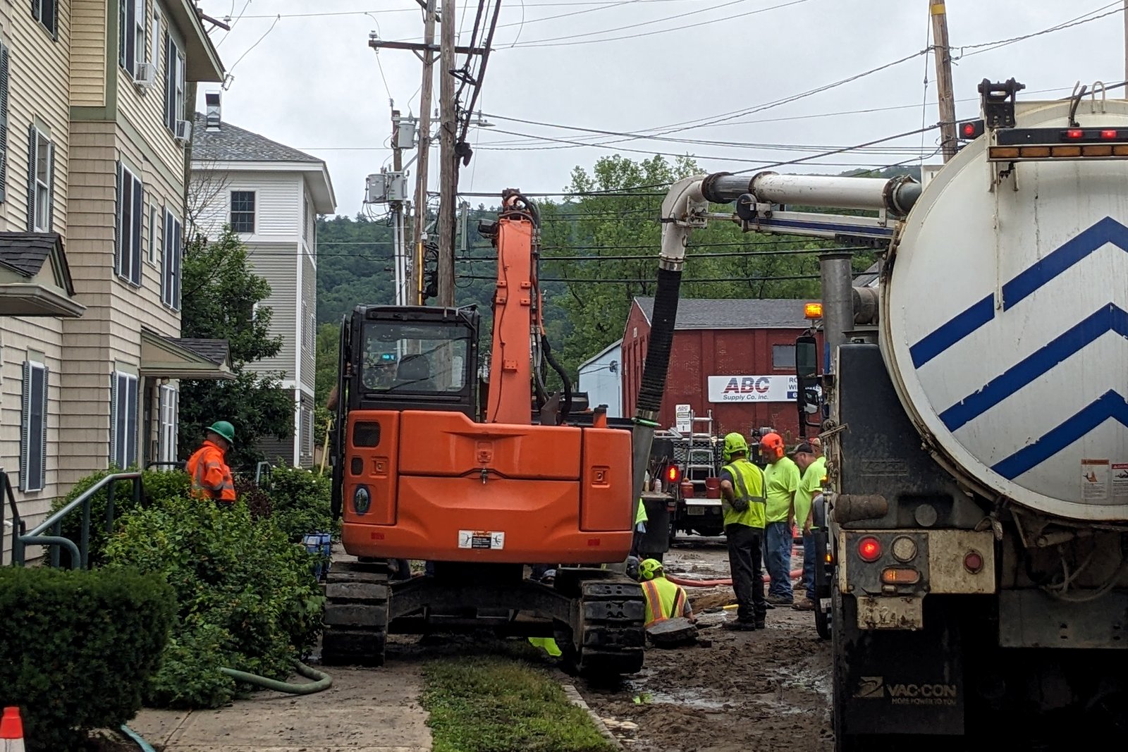 A street under construction with an orange excavator, a utility truck and workers in safety vests beside houses.
