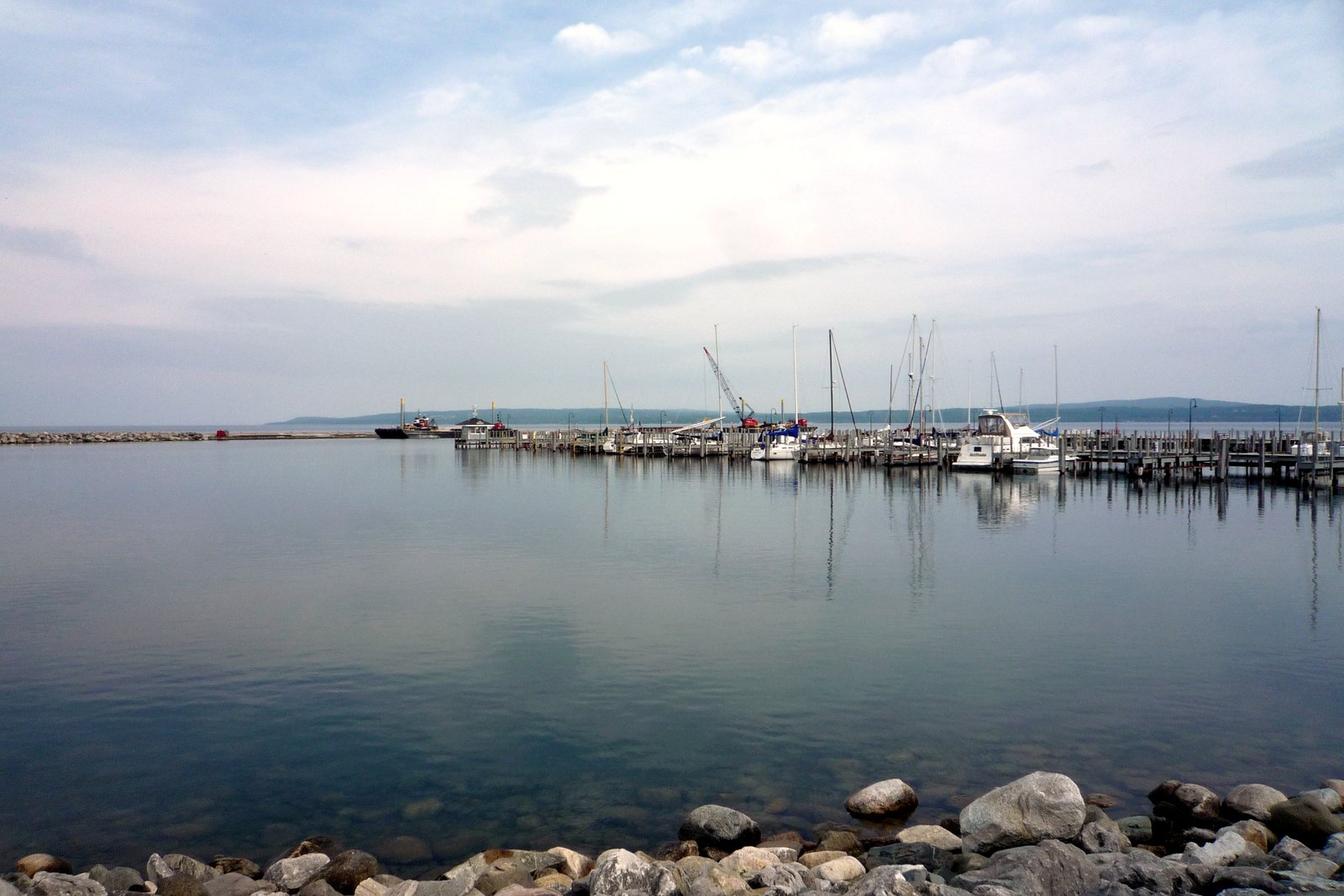 Calm bay water with a marina of docked boats in the distance and a rocky shoreline in the foreground.