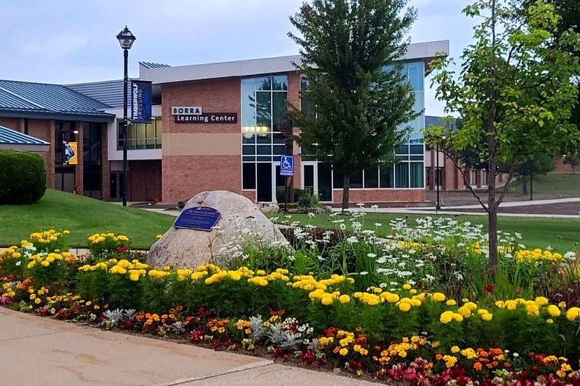 A brick and glass college building with a Borra Learning Center sign, behind a flower bed and a boulder with a plaque.