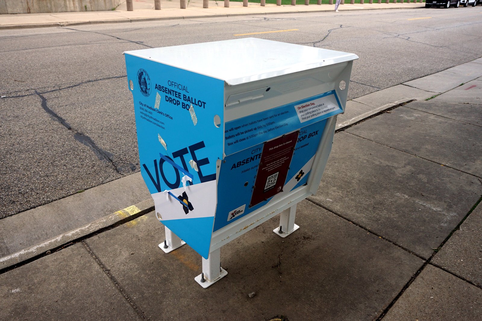 A blue official absentee ballot drop box on a sidewalk, with a white label on its front.