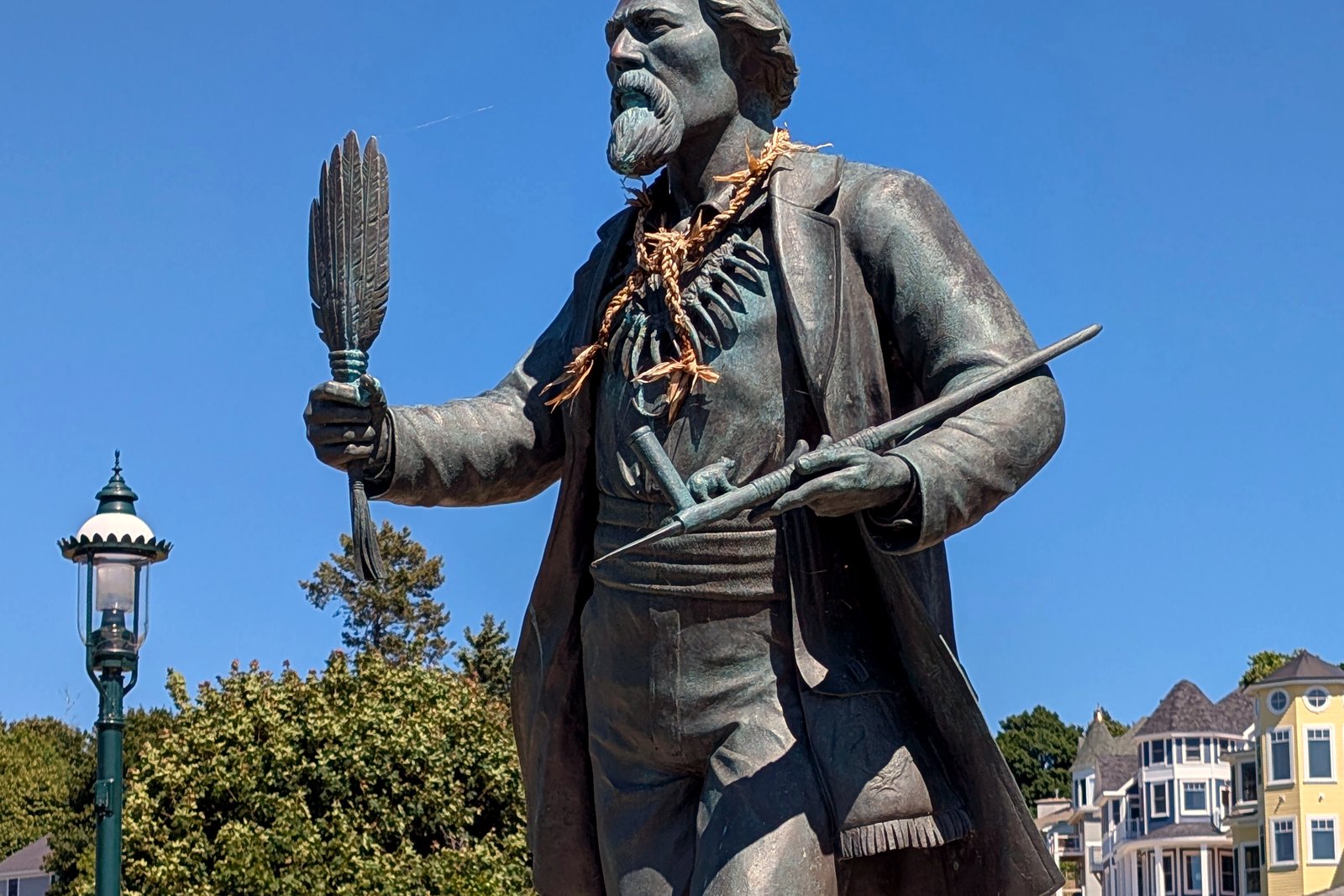 A bronze statue of a bearded man in a long coat holding a feathered staff, against a blue sky with a green lamppost and houses behind.