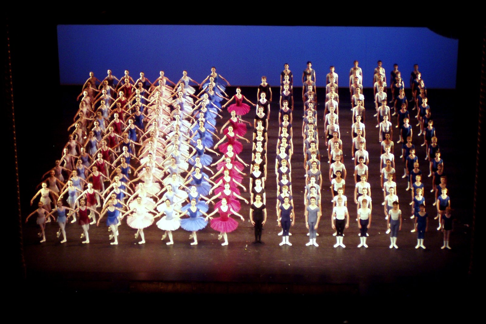 Rows of young ballet dancers in colorful tutus and practice clothes stand on a stage under blue light during a curtain call.