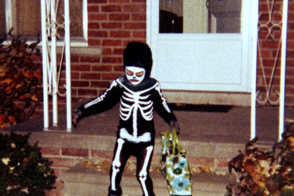 A small child in a skeleton costume stands on a front porch holding a trick-or-treat bag.